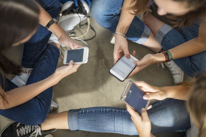 Drie jongeren (meisjes) zittend op de grond met mobiel in hun hand, ze hebben allen spijkerbroek aan en kijken op hun mobiel, je ziet ze van bovenaf